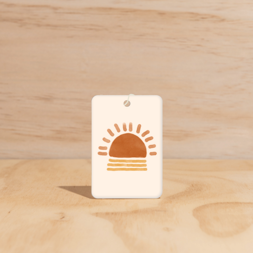 The Commonfolk air freshener, with a graphic of a sun on it, is displayed against a wooden background.