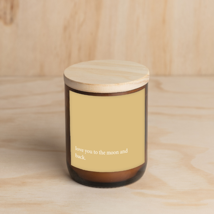 The Commonfolk scented candle with a yellow label that reads 'love you to the moon and back' in a white text. Wooden lid and amber jar.