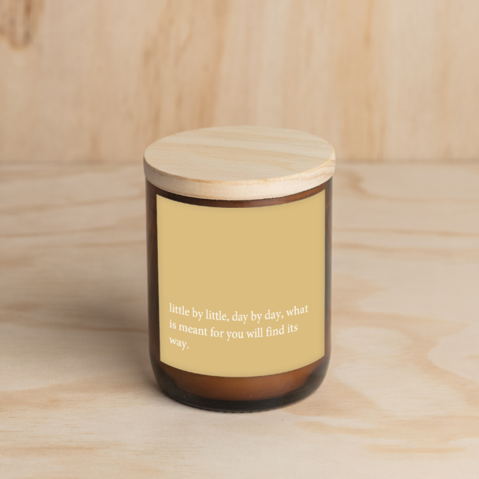 The Commonfolk scented brown jar candle with a wooden lid, features a yellow label with white text that reads 'little by little, day by day, what is meant for you will find its way.'.