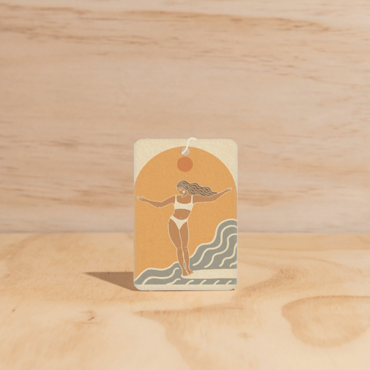The Commonfolk air freshener features a beige and orange color scheme with a graphic of a woman in a bikini with outstretched arms above a wave design.