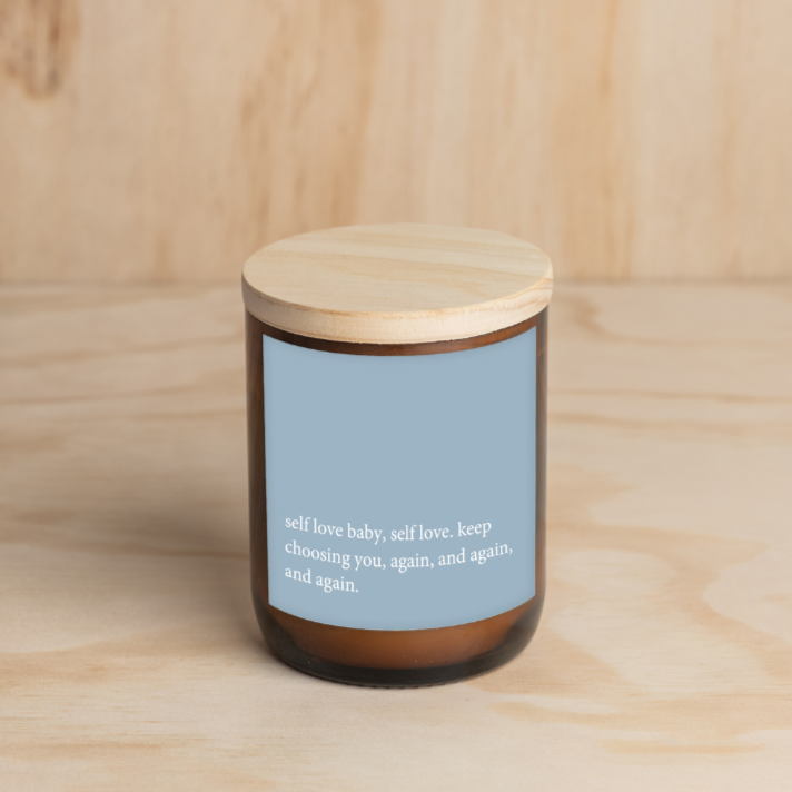 The Commonfolk scented amber jar candle with a wooden lid, features a blue label with white text that reads 'self love baby, self love, keep choosing you, again and again, and again.'.