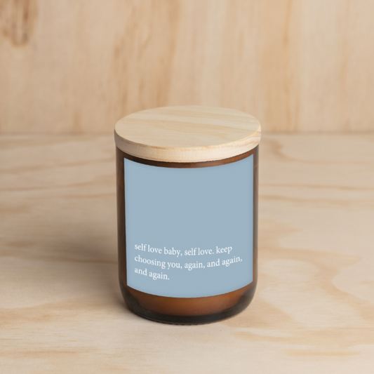 The Commonfolk scented amber jar candle with a wooden lid, features a blue label with white text that reads 'self love baby, self love, keep choosing you, again and again, and again.'.