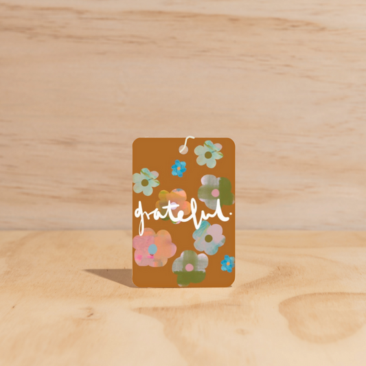 The Commonfolk air freshener, with a floral design and the word 'grateful' written across the center, is displayed on a wooden surface.