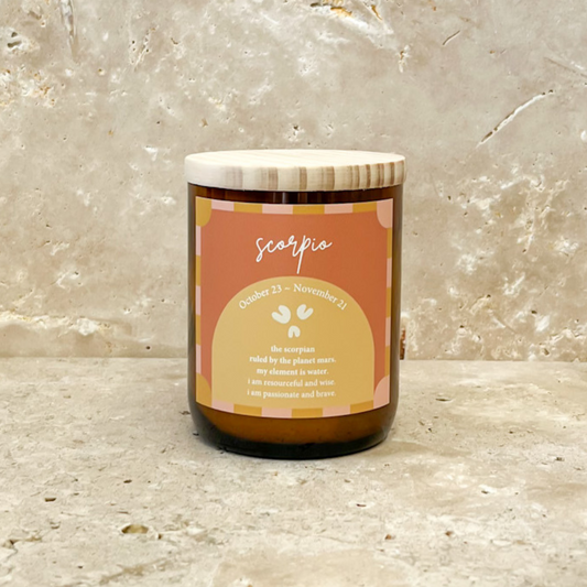 The Commonfolk scented soy wax candle with a wooden lid, in a brown glass jar, labeled 'Scorpio' with an orange and yellow label, on a textured background.
