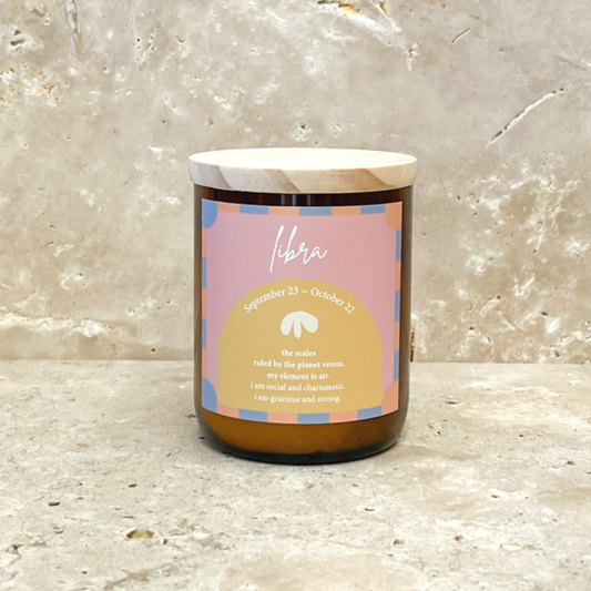 The Commonfolk scented soy wax candle with a wooden lid, in an amber glass jar, labeled 'Libra' with a pink and yellow color scheme, on a marble surface.