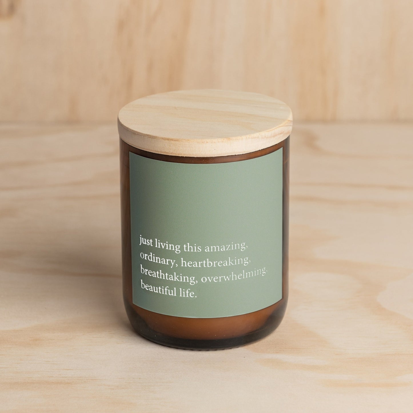 The Commonfolk scented soy candle with a wooden lid and a green label featuring a quote is placed on a wooden surface.