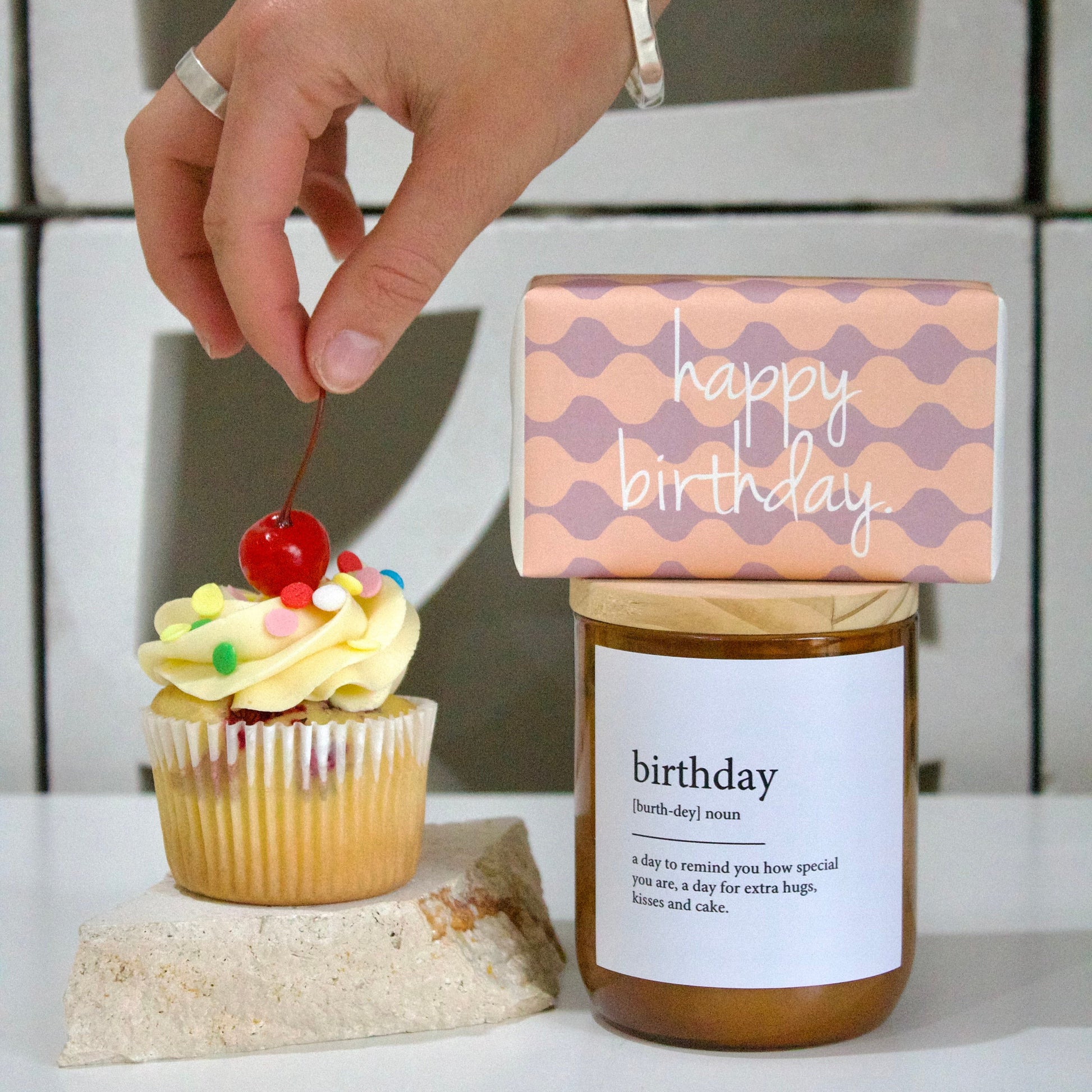 Cupcake with cherry and candle, 'happy birthday' card, and jar labeled 'birthday' on a white surface.