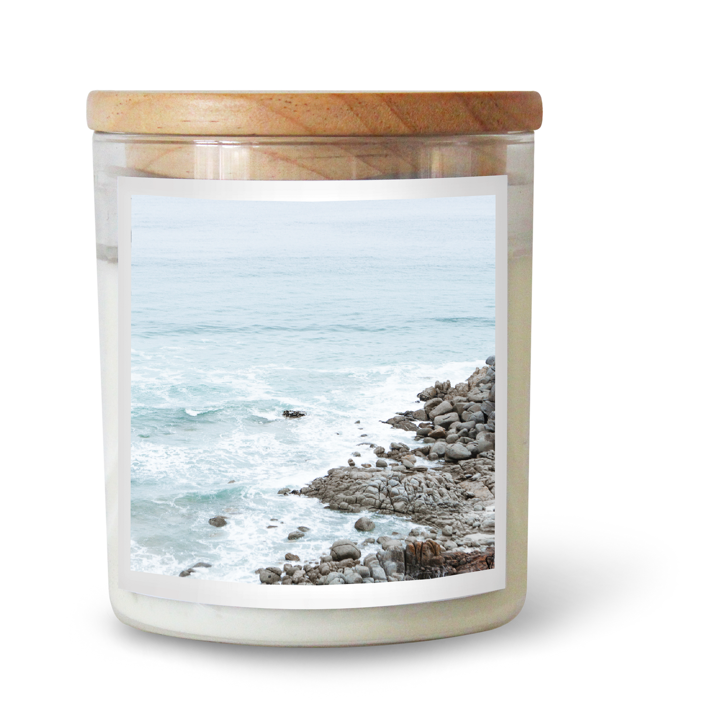 The Commonfolk scented clear glass jar candle with a wooden lid and a label depicting a beach scene with rocks and the ocean.