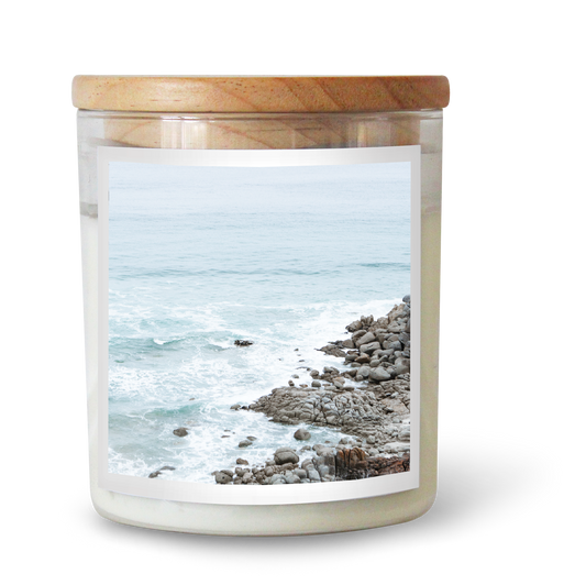 The Commonfolk scented clear glass jar candle with a wooden lid and a label depicting a beach scene with rocks and the ocean.