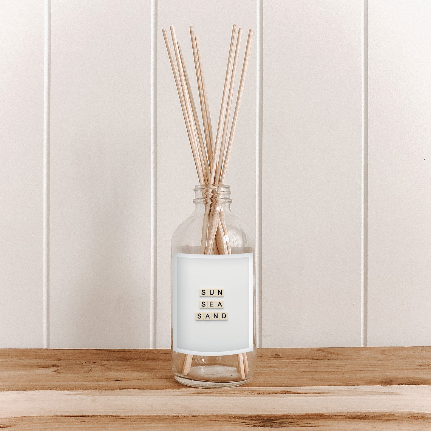 The Commonfolk room diffuser with reeds labeled 'Sun Sea Sand' on a wooden surface with a white paneled background