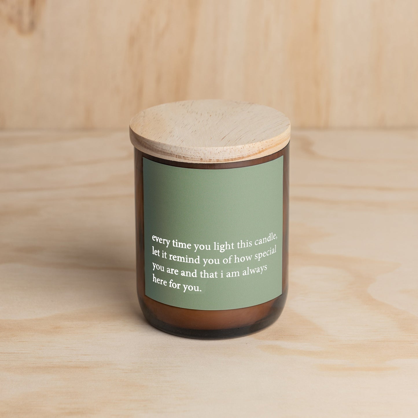 The Commonfolk scented candle, with a green label that contains a heartfelt quote, is placed in an amber jar with a wooden lid, set against a wooden background.