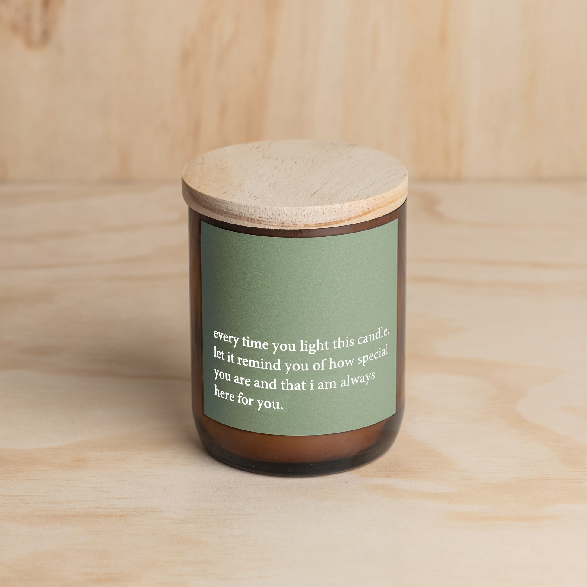 The Commonfolk scented candle, with a green label that contains a heartfelt quote, is placed in an amber jar with a wooden lid, set against a wooden background.