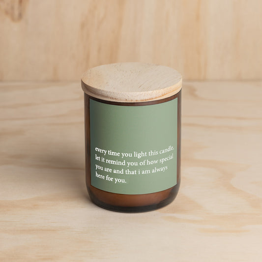 The Commonfolk scented candle, with a green label that contains a heartfelt quote, is placed in an amber jar with a wooden lid, set against a wooden background.