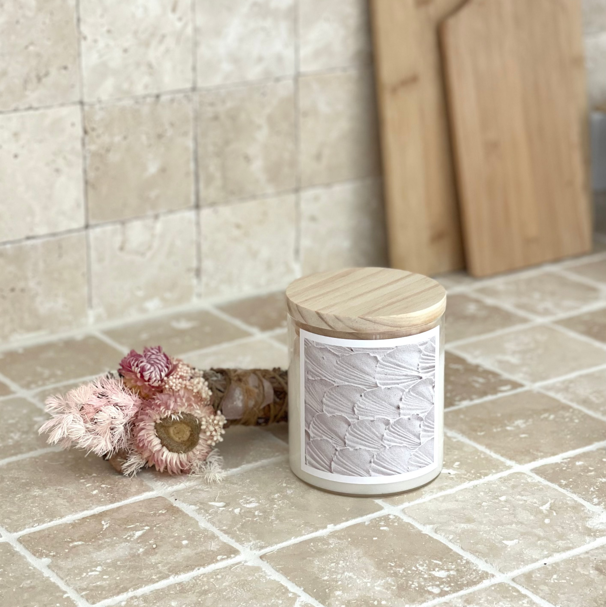White candle with wooden lid on a tiled floor next to a wooden cutting board and dried flowers.