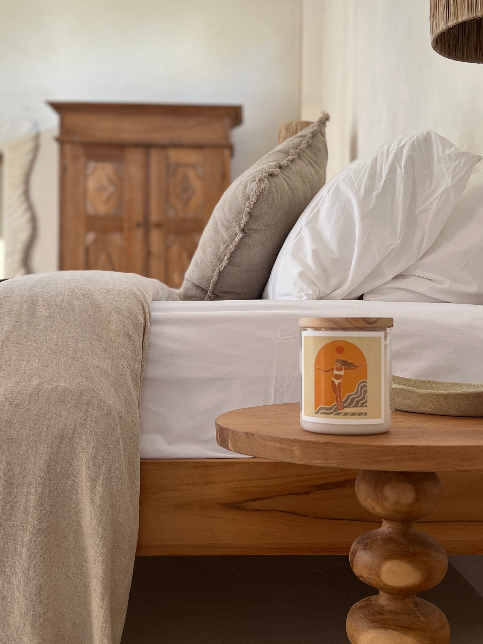 A wooden nightstand with a scented candle and a white bed in the background.