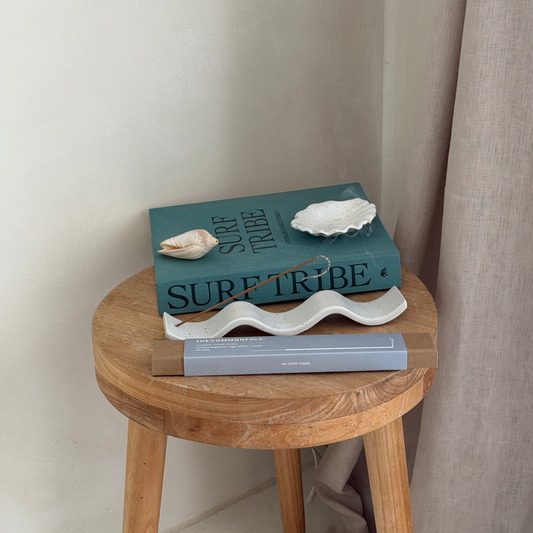 A teal hardcover book titled 'SURF TRIBE' styled on a round wooden stool next to a beige curtain, with decorative seashells on top with incense burning in incense holder.