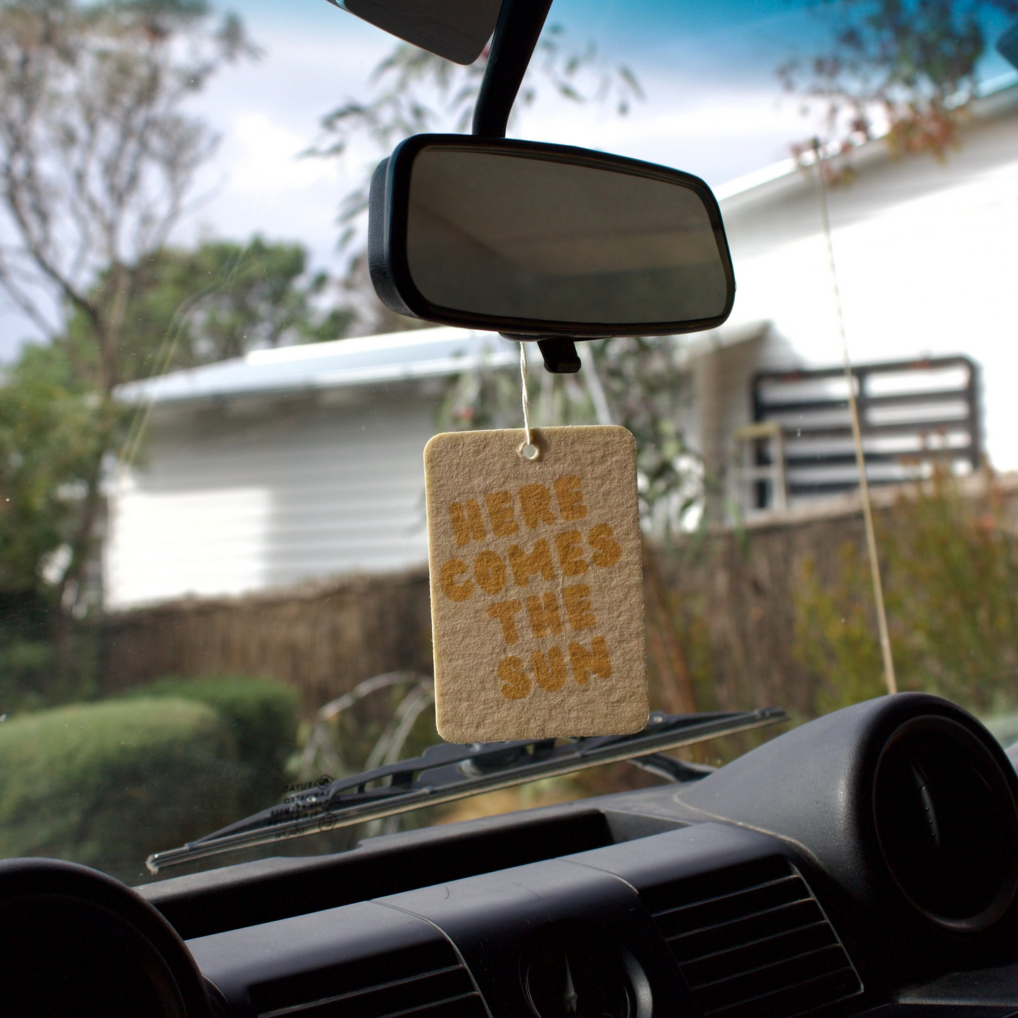 Car air freshener hanging from the rearview mirror with 'Here Comes the Sun' text.