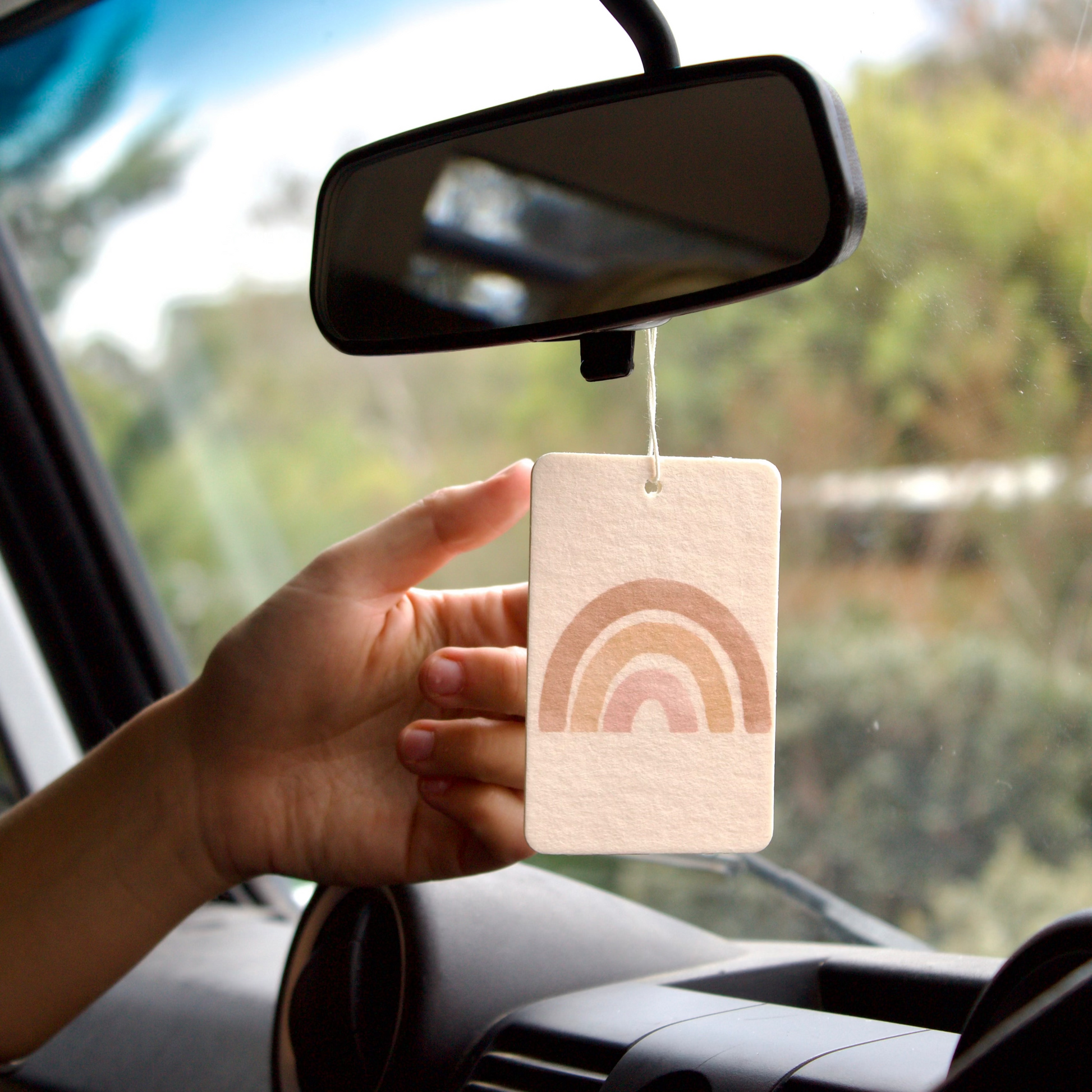 Hand holding a car air freshener with rainbow design in front of a car mirror.
