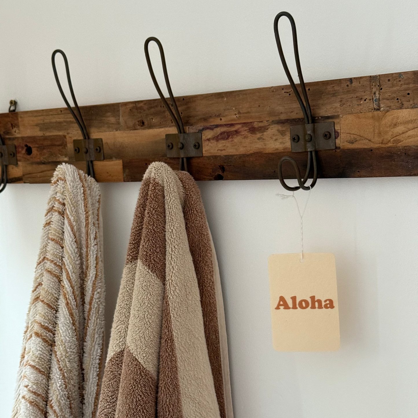 Wooden towel rack with towels and an air freshener 'Aloha' sign on a white wall.