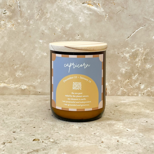 The Commonfolk scented Capricorn-themed candle, with a label in shades of yellow and blue, a wooden lid, and an overall cozy appearance, is placed against a textured background.