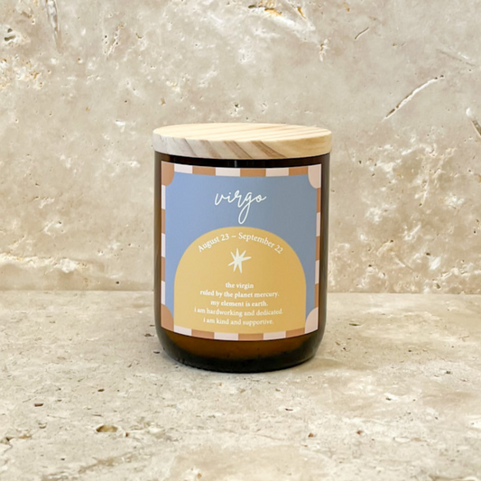 The Commonfolk scented soy wax candle in an amber jar with a wooden lid, labeled 'Virgo' with a yellow and blue color scheme, is set against a marble background.