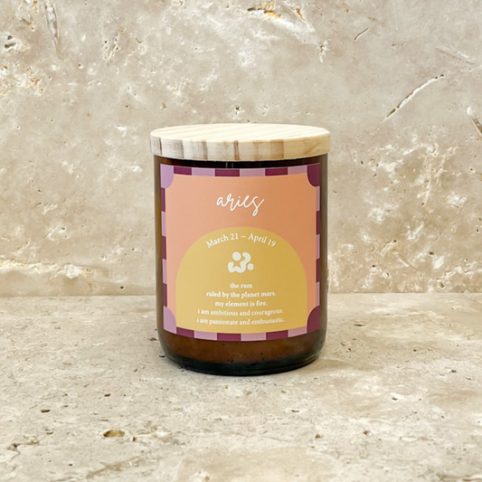 The Commonfolk scented soy wax candle is in a brown jar with a label that has 'Aries' written on it, alongside a date range and a brief description. The candle is placed against a textured marble background.