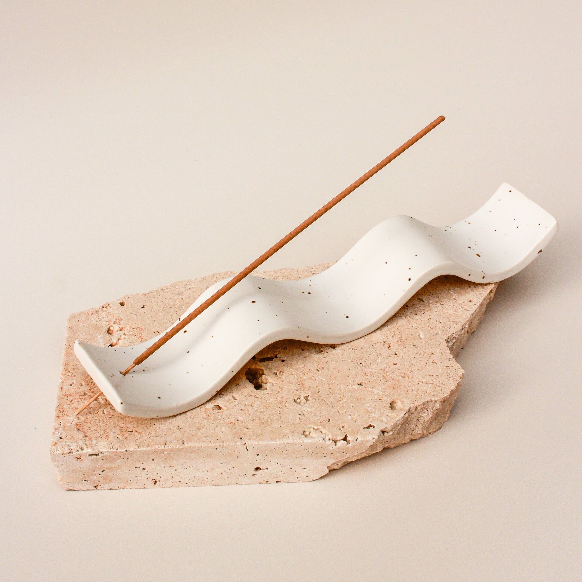 A beige ceramic incense holder from The Commonfolk with brown speckles and an incense stick placed on top, on a neutral background.