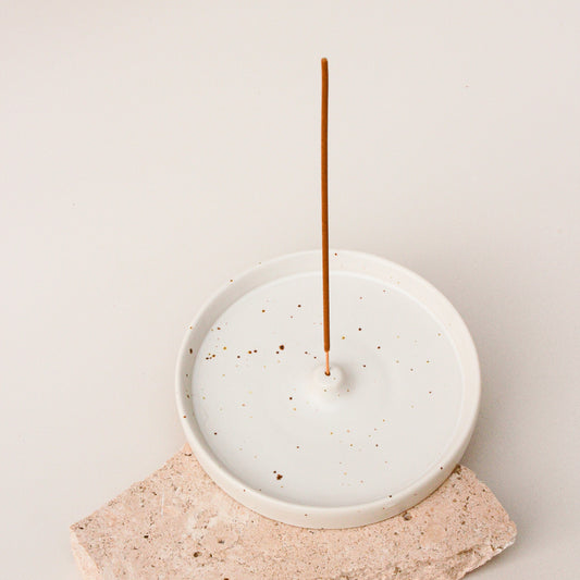 A white ceramic incense holder from The Commonfolk with brown speckles on a neutral background, displaying a single incense stick placed on top.