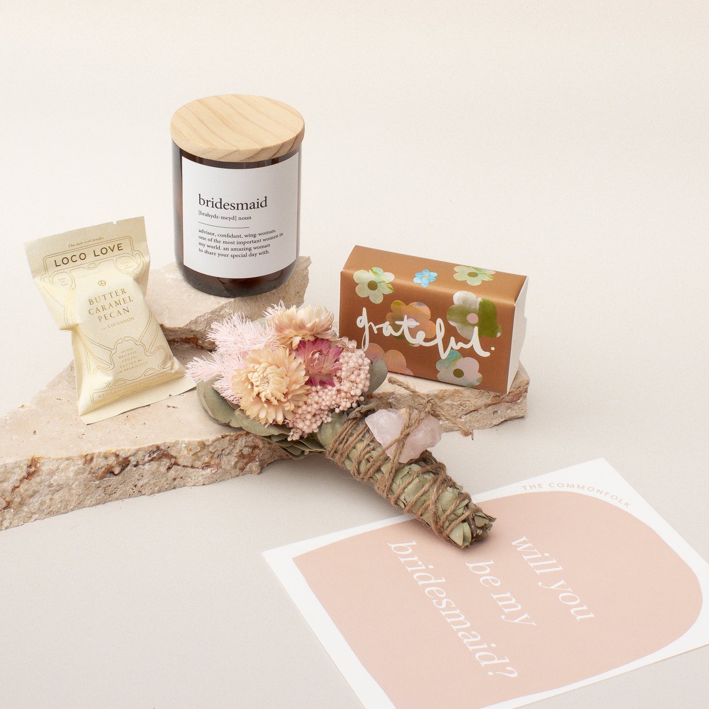 The Commonfolk gift box for bridesmaids features a midi candle, chocolate, a body bar, a dried flowers smudge stick with a crystal, and a gift card, all displayed on a wooden surface.