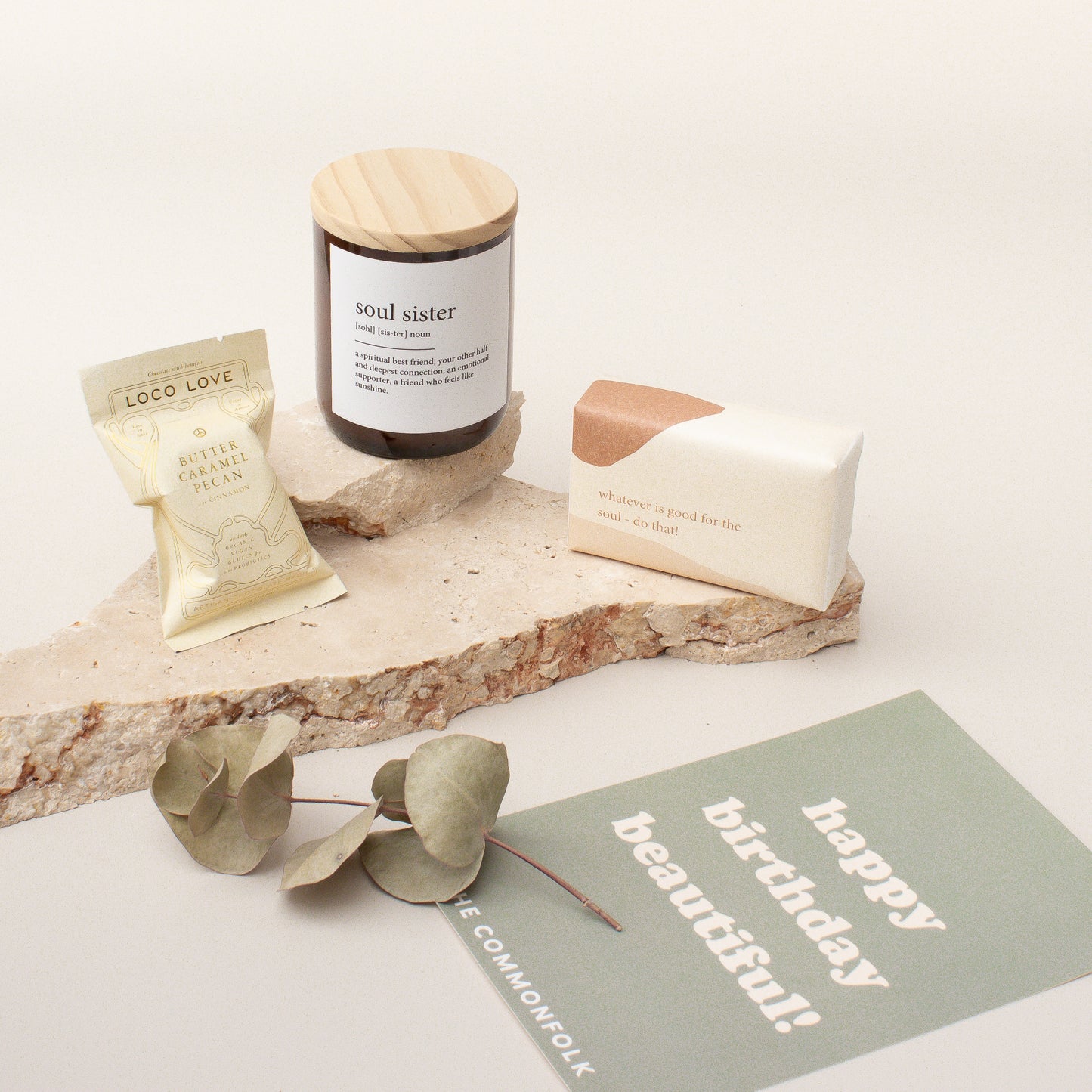 The Commonfolk gift box arrangement includes a midi candle, chocolate, body bar, and a gift card, presented on a stone surface with decorative greenery and a 'Happy Birthday' card.