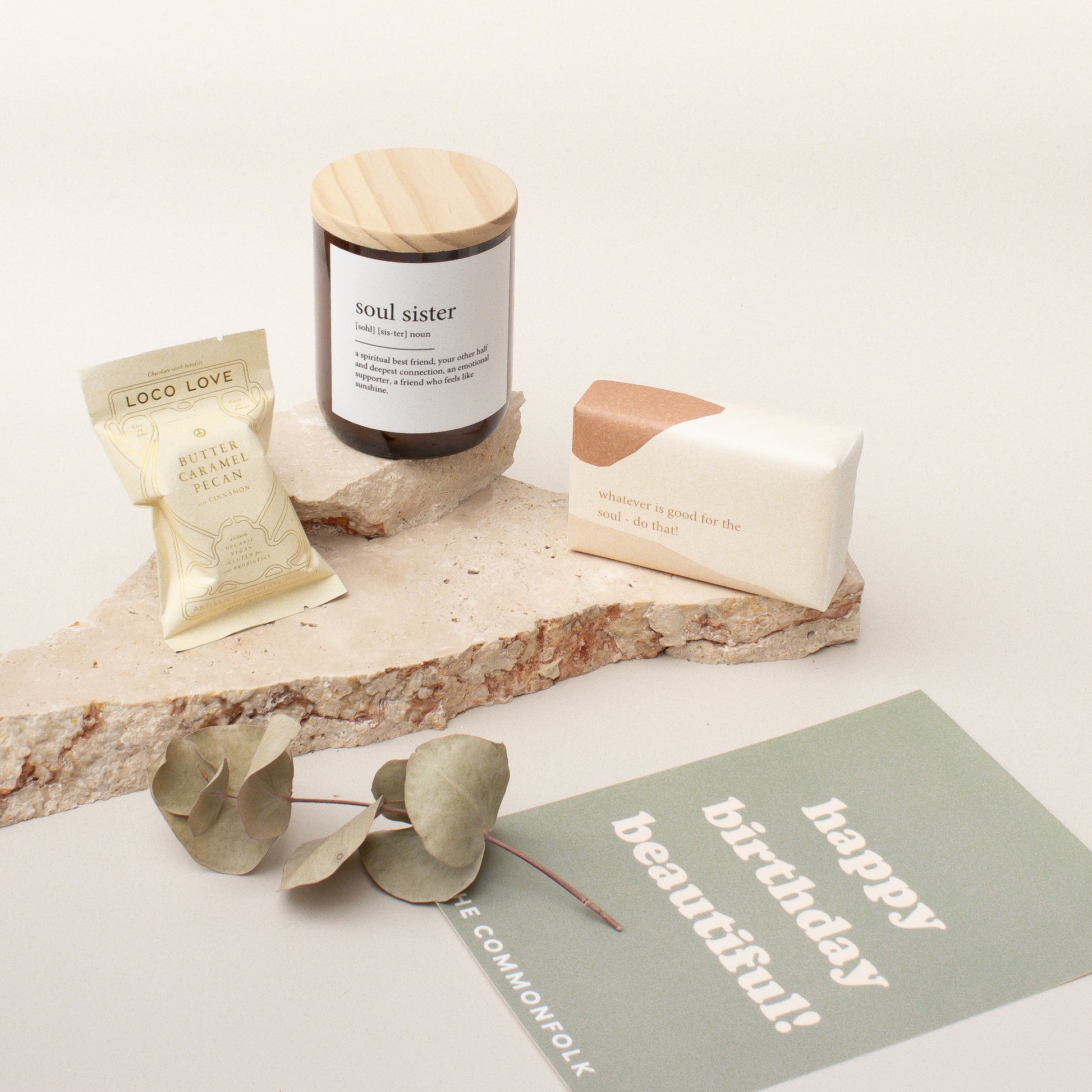 The Commonfolk gift box arrangement includes a midi candle, chocolate, body bar, and a gift card, presented on a stone surface with decorative greenery and a 'Happy Birthday' card.