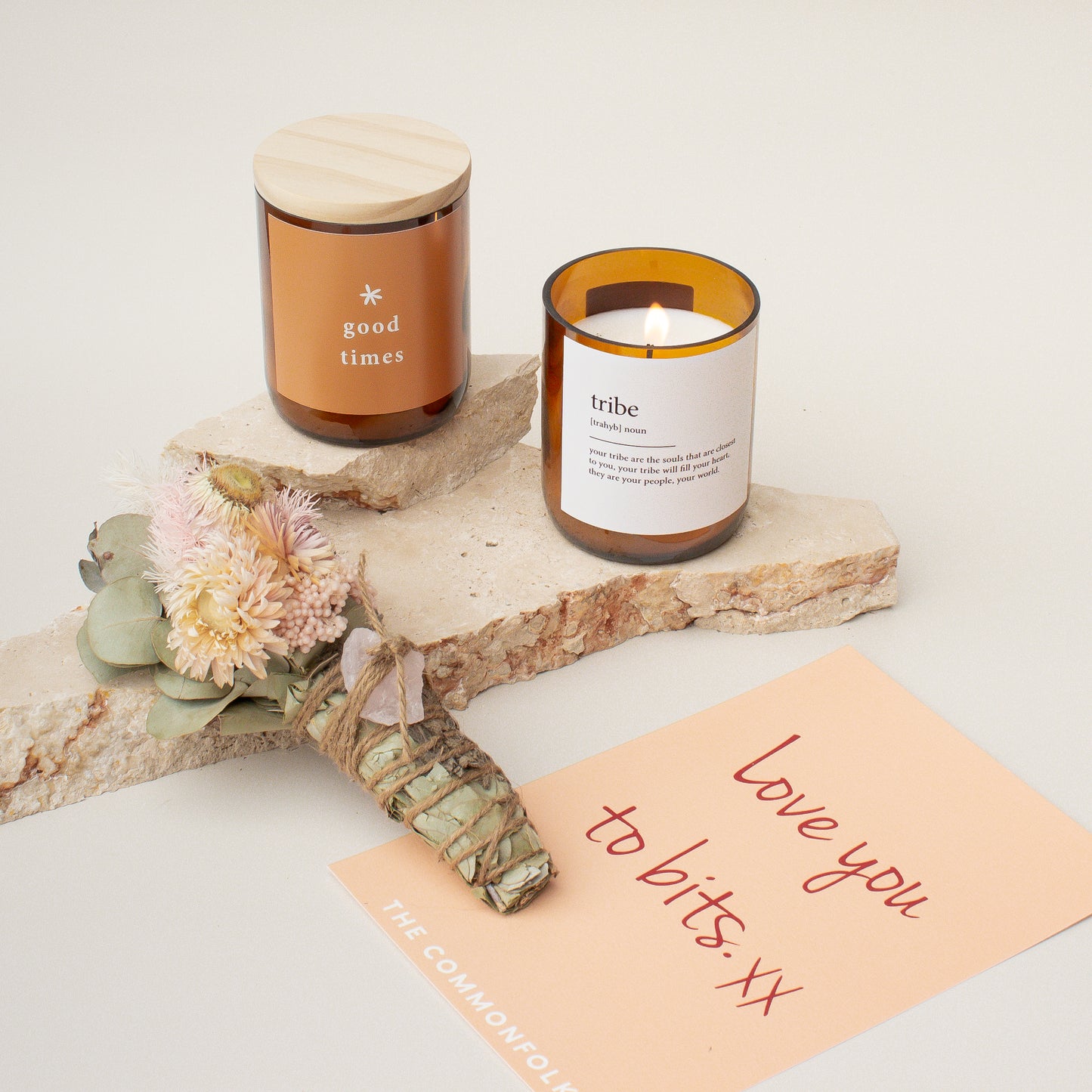 The Commonfolk gift set includes two candles in glass jars with wooden lids and a gift card placed on a natural-looking surface. It is accompanied by a dried flower smudge stick with a rose quartz crystal.