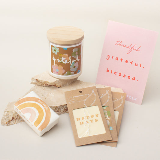 The Commonfolk gift box collection includes a candle with a floral design on the container, a body bar with a rainbow design, air fresheners, and a gift card with written messages. All items are presented on a neutral background.