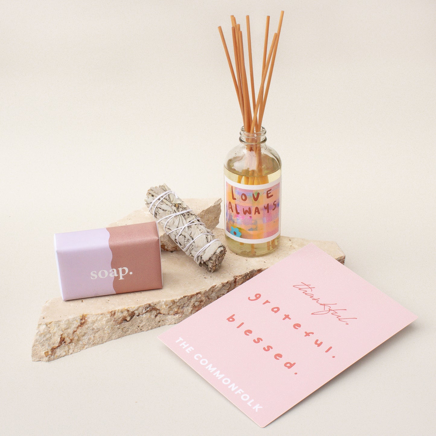 The Commonfolk gift box presentation features a room diffuser, a dried sage smudge stick, a body bar, and a gift card, all laid out on a stone surface with a pink card.