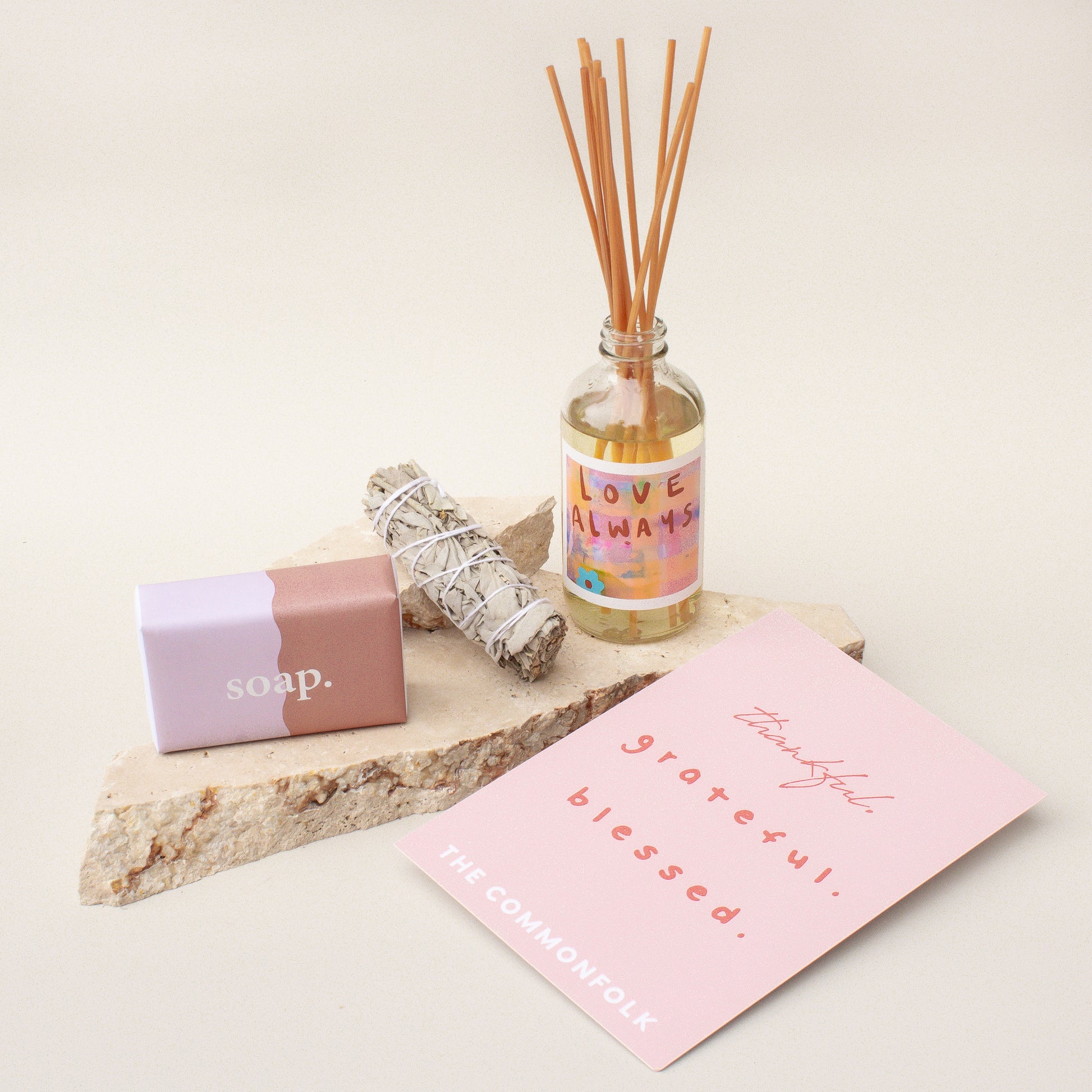 The Commonfolk gift box presentation features a room diffuser, a dried sage smudge stick, a body bar, and a gift card, all laid out on a stone surface with a pink card.