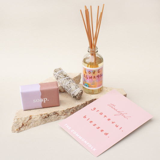 The Commonfolk gift box presentation features a room diffuser, a dried sage smudge stick, a body bar, and a gift card, all laid out on a stone surface with a pink card.