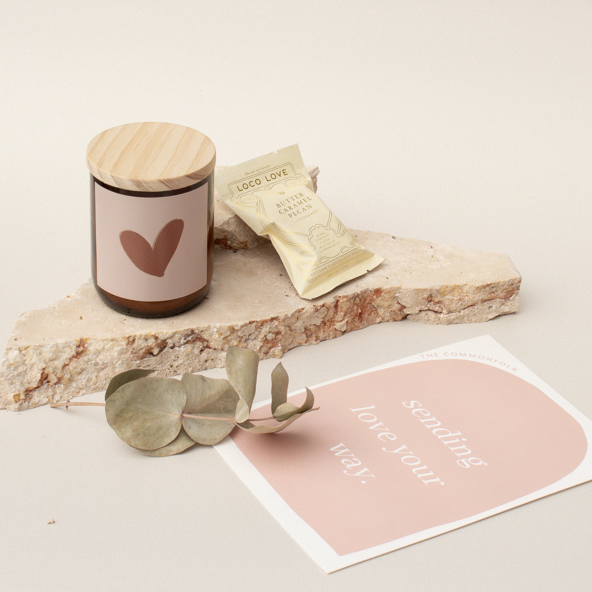 The Commonfolk gift box is set up with a candle, chocolate, and a gift card, presented on a stone surface with a eucalyptus leaf and a pink note card.