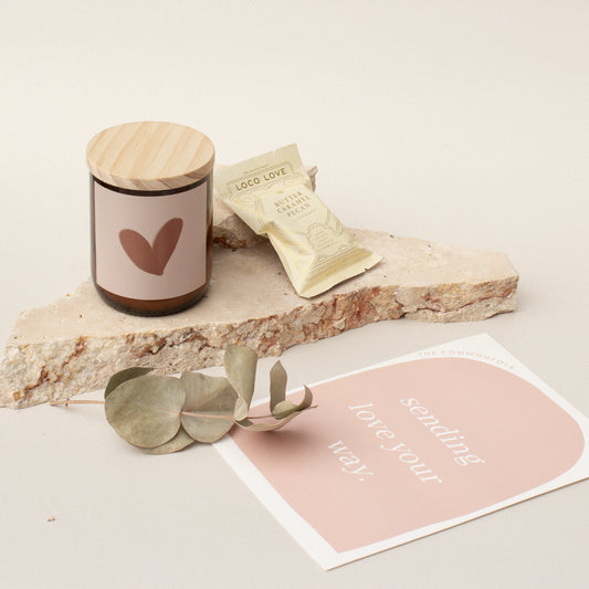The Commonfolk gift box is set up with a candle, chocolate, and a gift card, presented on a stone surface with a eucalyptus leaf and a pink note card.