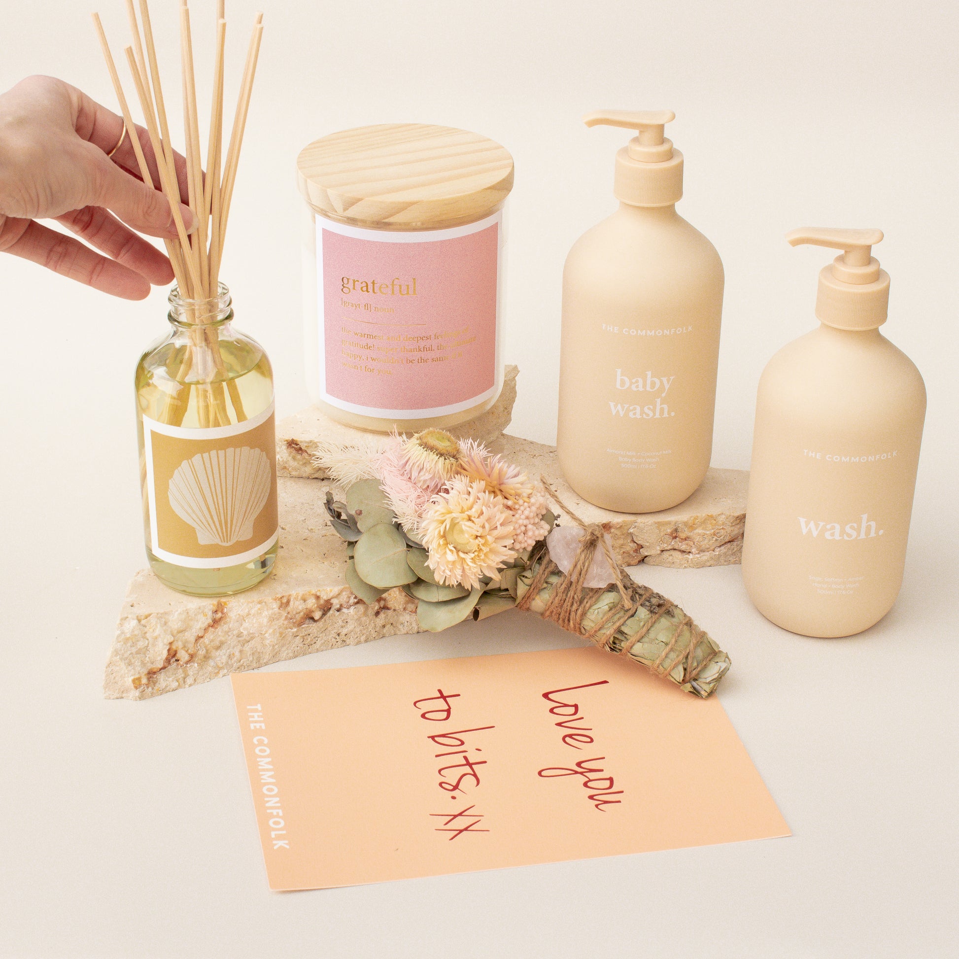 The Commonfolk gift set, including a candle, wash, lotion, diffuser, and a gift card, is arranged on a stone with a person's hand placing a reed into the diffuser.