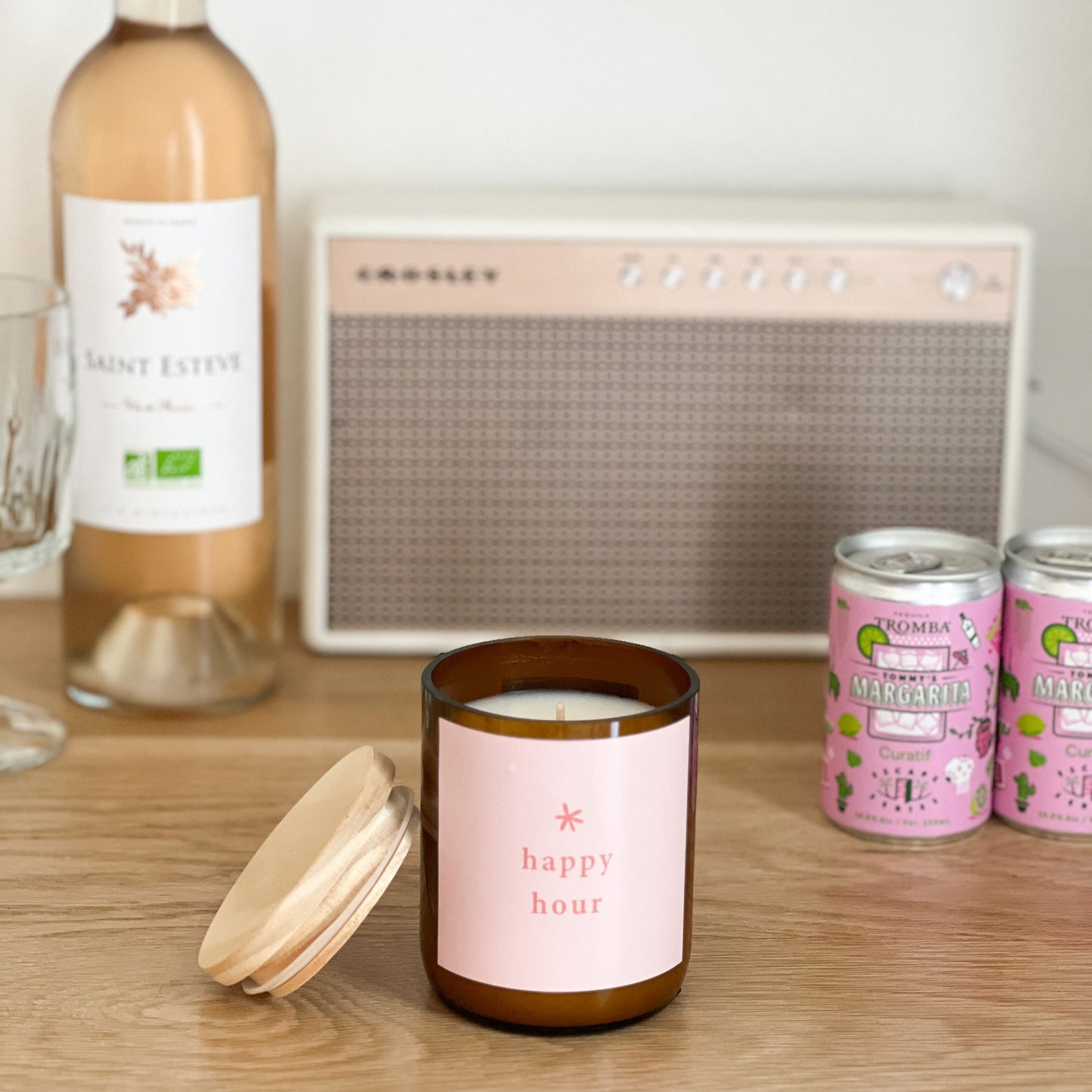 Candle with 'happy hour' label on a table with wine, cans, and radio in the background