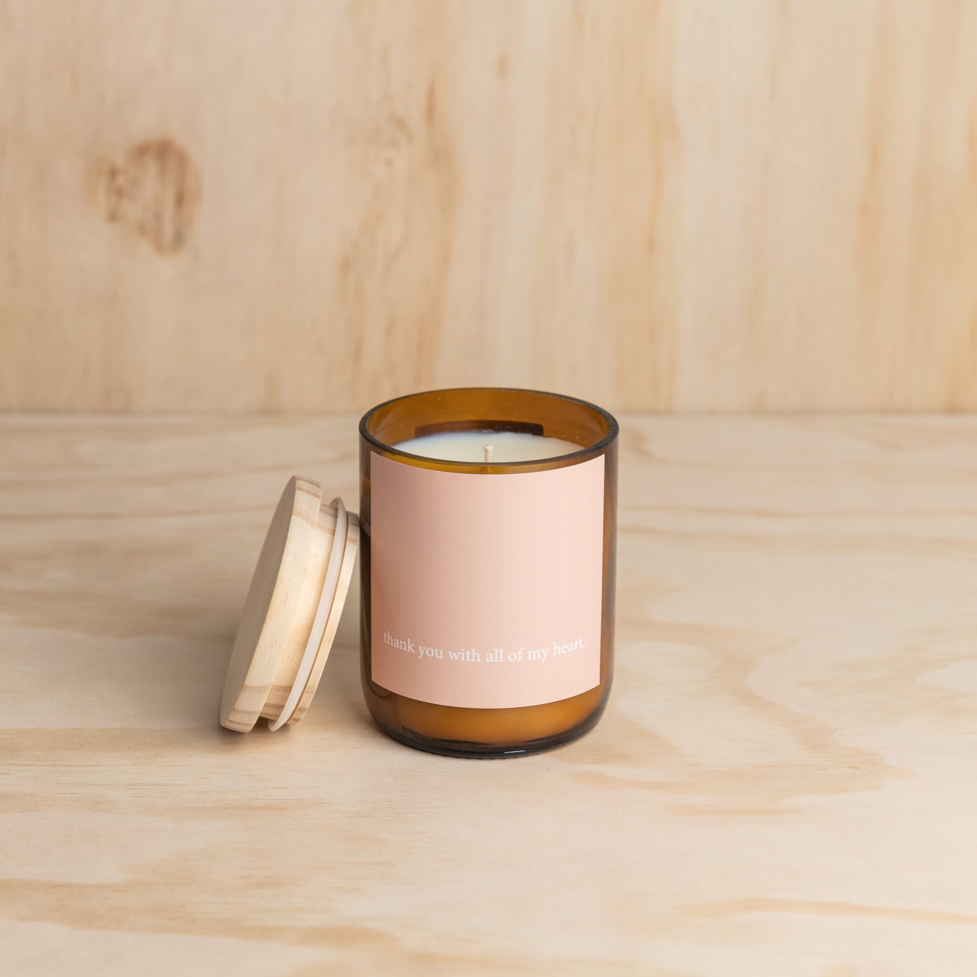 A candle with a wooden lid and a pink label, placed on a neutral background.