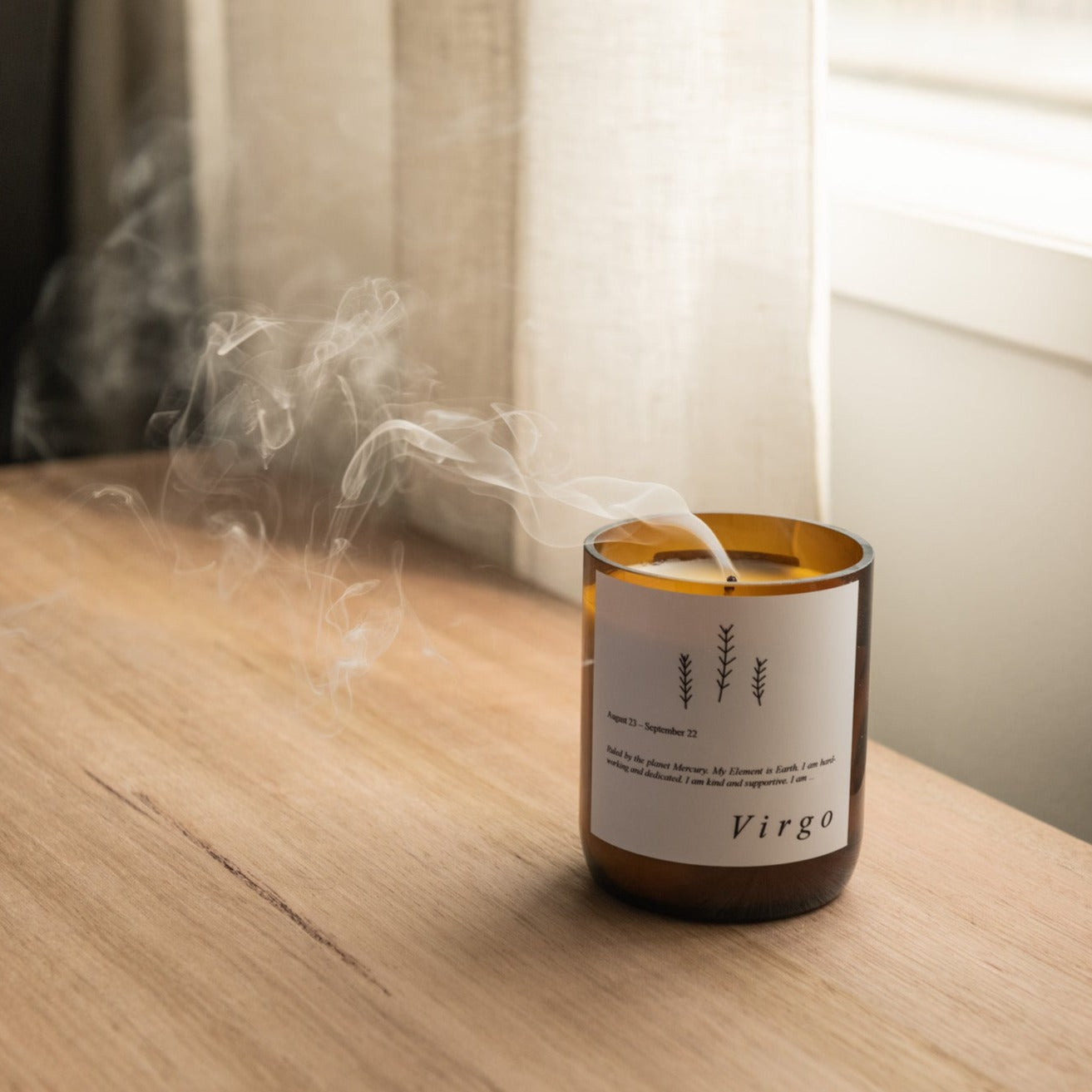 The Commonfolk scented soy wax candle with a label that reads 'Virgo', in an amber glass jar, with smoke rising from the top, placed on a wooden surface.