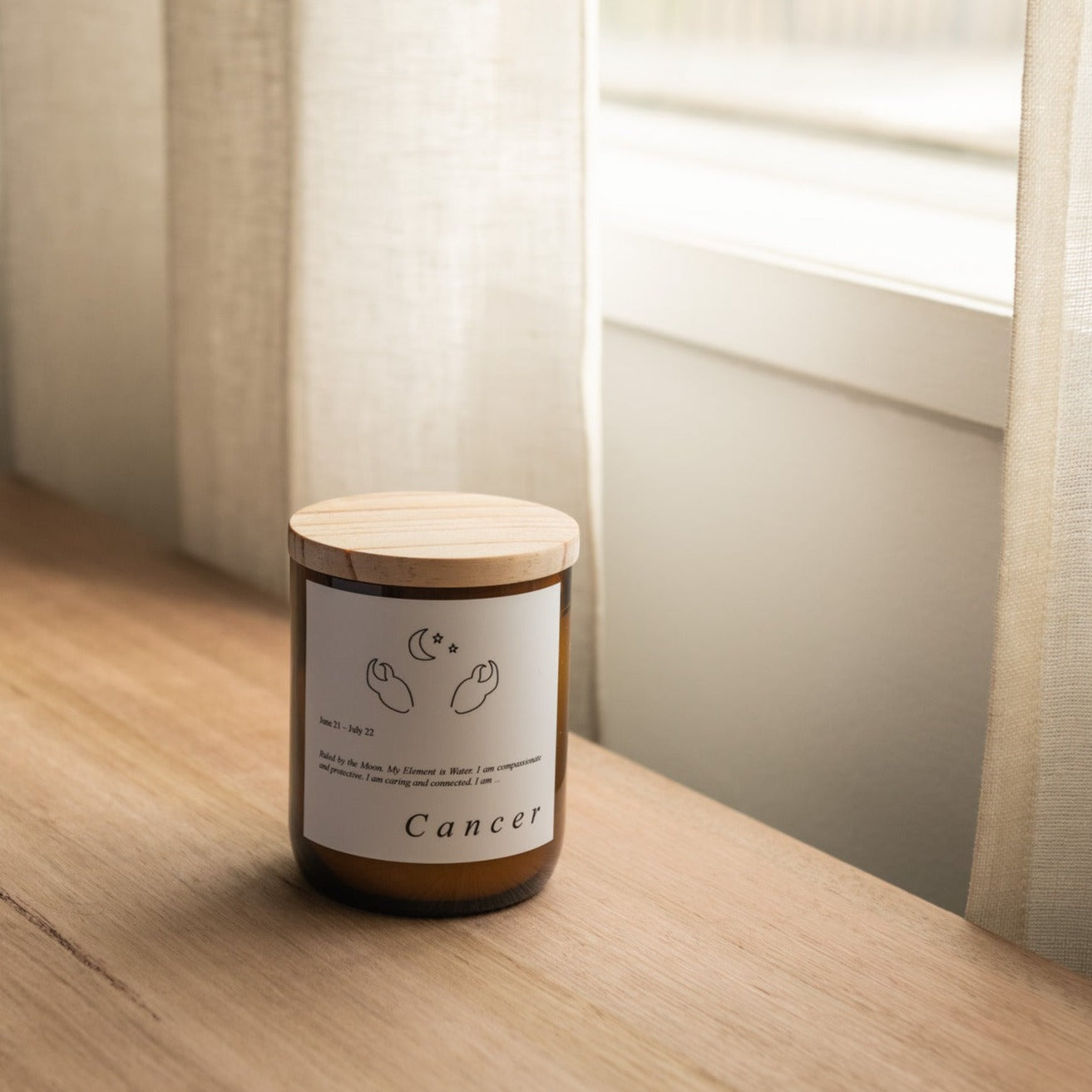 The Commonfolk scented candle with a wooden lid, labeled 'Cancer', placed on a wooden surface, with a window and curtains in the background.