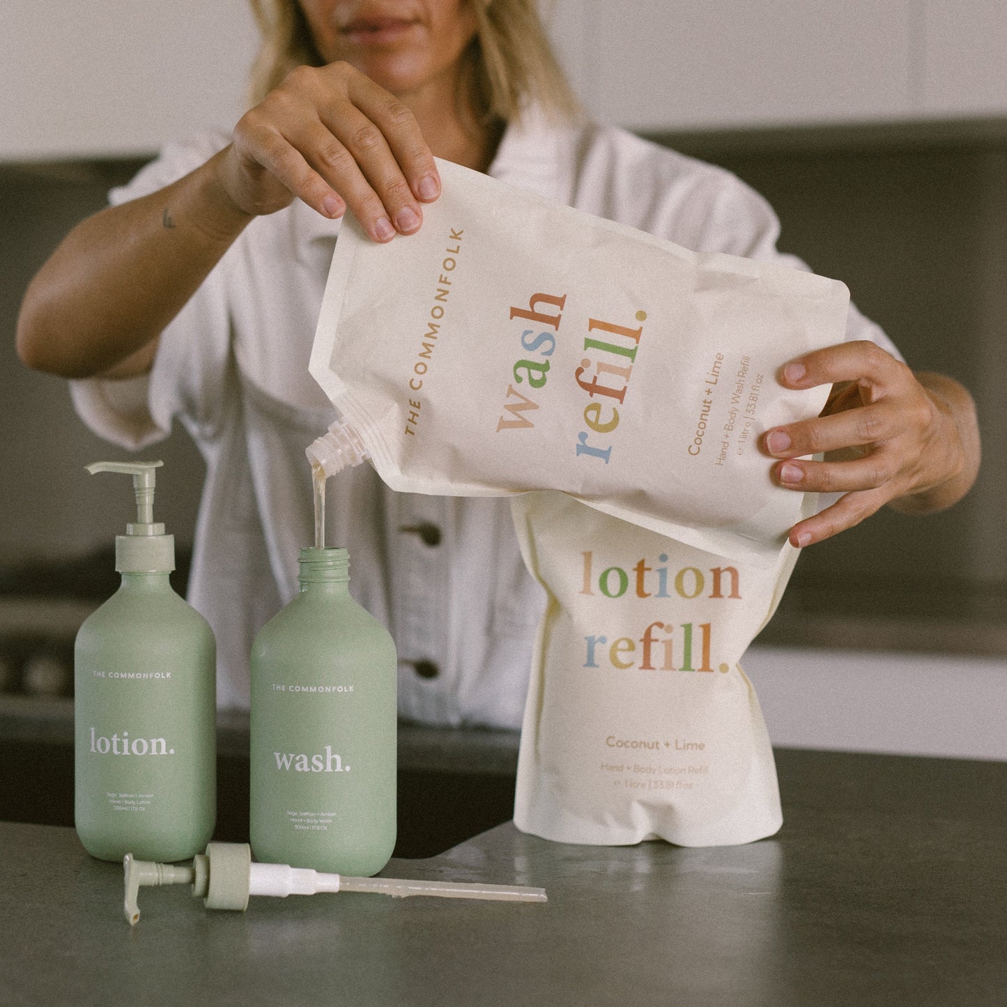The Commonfolk home starter pack. A person is pouring liquid from a refill pouch into a bottle, with another bottle alongside. The pouch has the text 'wash refill' printed on it.