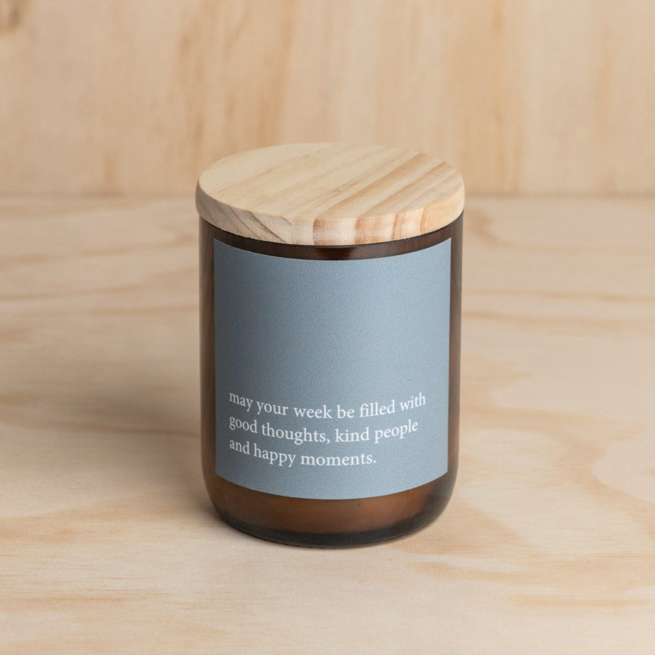 The Commonfolk scented soy candle in an amber jar with a wooden lid, features a blue label with white text that reads 'May your week be filled with good thoughts, kind people and happy moments.'.