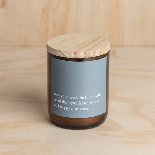 The Commonfolk scented soy candle in an amber jar with a wooden lid, features a blue label with white text that reads 'May your week be filled with good thoughts, kind people and happy moments.'.