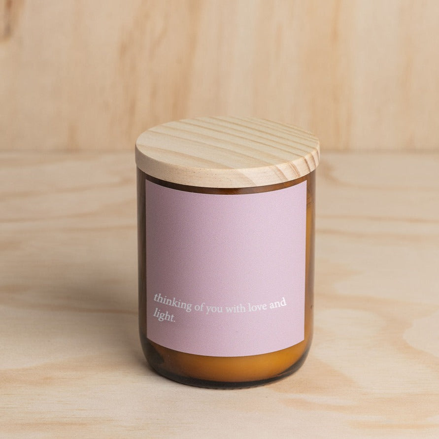 The Commonfolk scented mid-sized amber glass candle jar with a wooden lid and a purple label that reads 'thinking of you with love and light.'