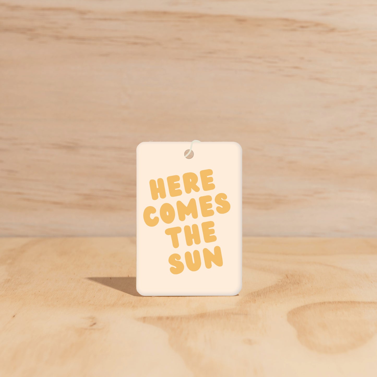 The Commonfolk air freshener with the text 'HERE COMES THE SUN' printed in yellow on a white background, displayed against a wooden backdrop.