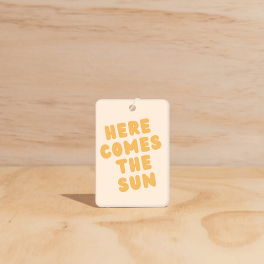 The Commonfolk air freshener with the text 'HERE COMES THE SUN' printed in yellow on a white background, displayed against a wooden backdrop.