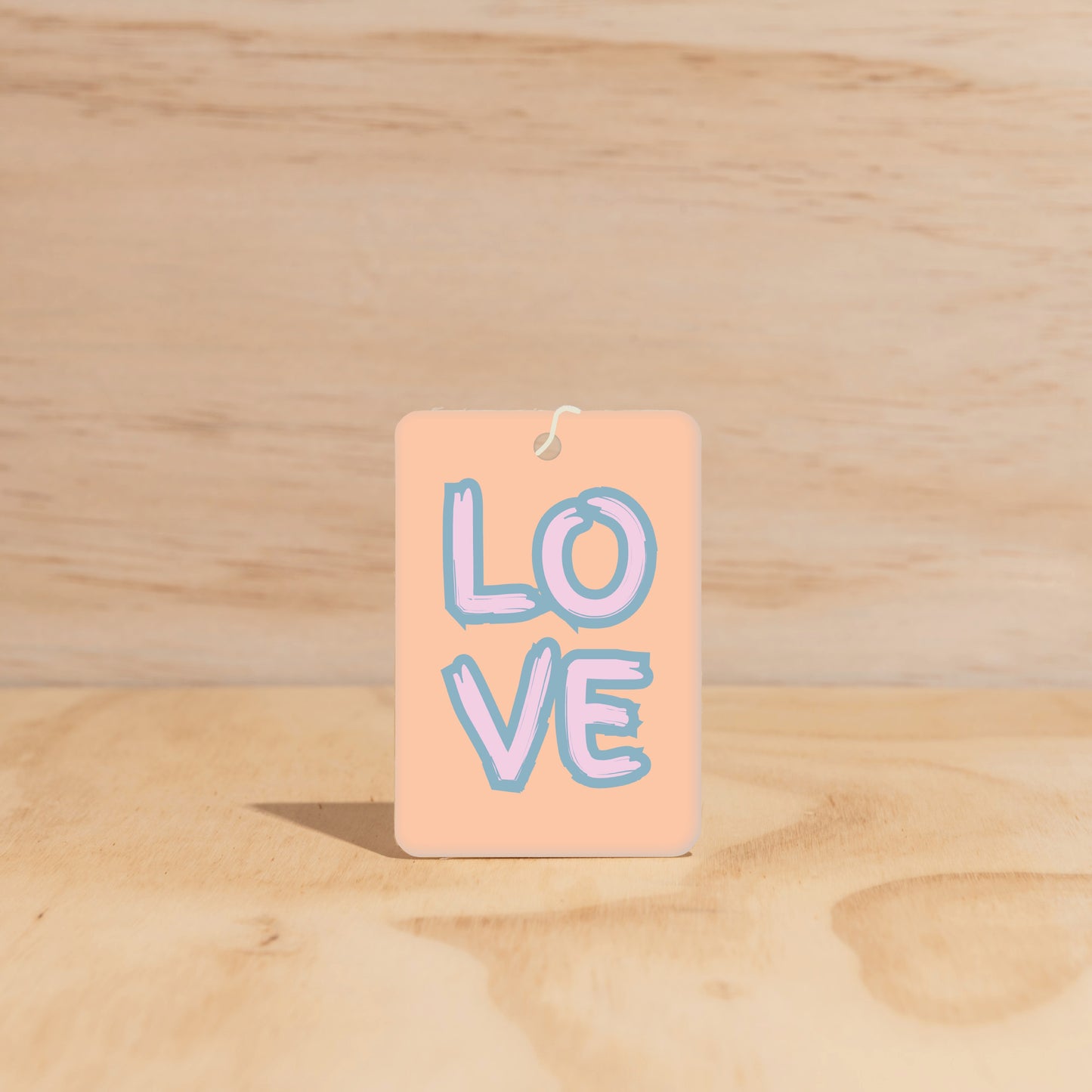 The Commonfolk air freshener with 'LOVE' written in blue on a beige background, hanging against a wooden backdrop.